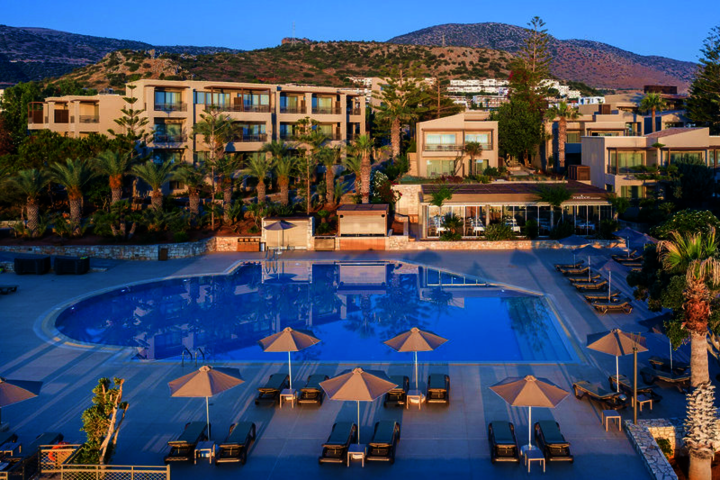 Großer Pool mit Liegen und Sonnenschirmen vor mehrstöckigen Hotelgebäuden und Palmen unter klarem Himmel.