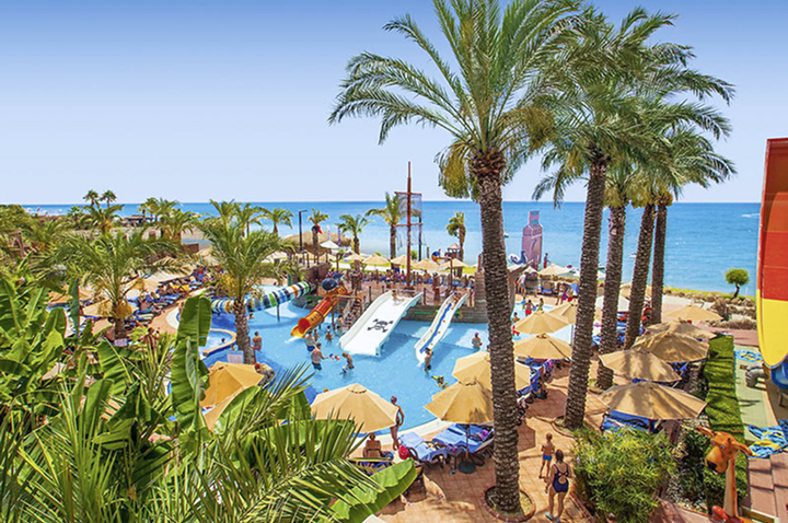 Ein großer Pool mit einer Wasserrutsche, umgeben von Palmen, strahlt Urlaubsstimmung und Entspannung aus.