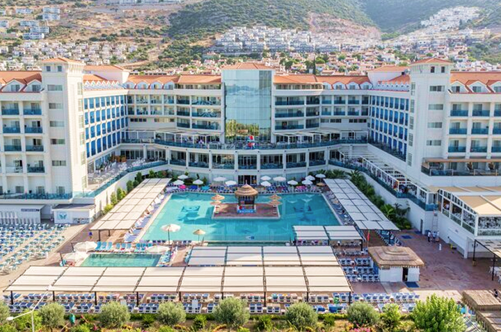 Großes Hotelgebäude mit mehreren Stockwerken um einen zentralen Poolbereich mit Sonnenschirmen und Liegen.