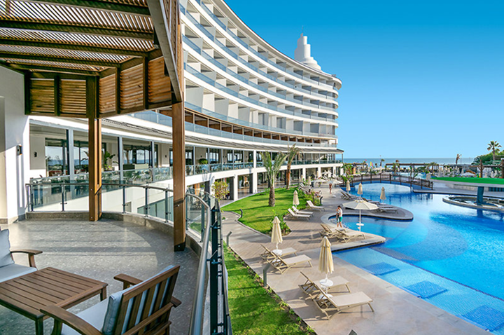 Gebogenes mehrstöckiges Hotelgebäude mit Glasfronten und großem Außenpool mit Liegestühlen und Sonnenschirmen