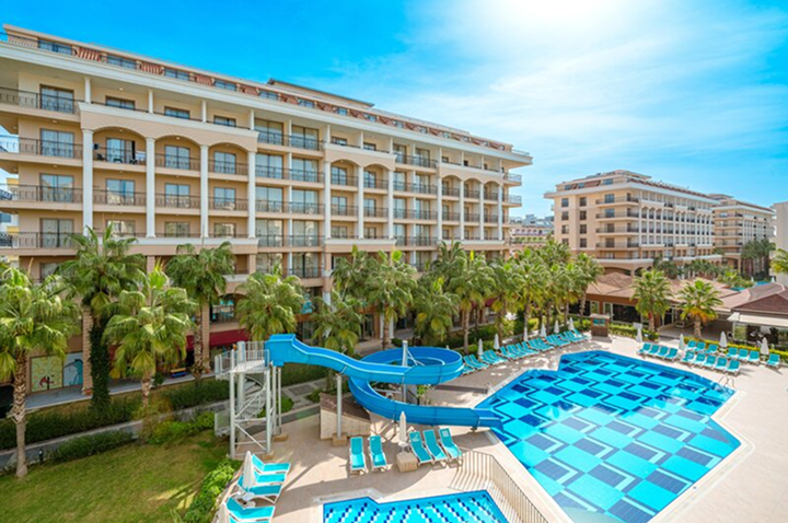 Großes Hotelgebäude mit mehreren Balkonen, davor ein Pool mit blauen Fliesen, Wasserrutsche und Liegestühlen, umgeben von Palmen.
