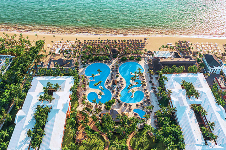 Luftaufnahme eines Resorts mit zwei großen, geschwungenen Pools, umgeben von Palmen und weißen Gebäuden am Strand mit türkisfarbenem Meer.