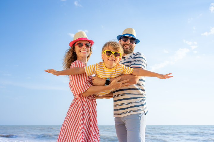 Familie am Strand: Kind mit gelb-weiß gestreiftem T-Shirt wird von Eltern gehalten, Mutter trägt rot-weiß gestreiftes Kleid, Vater blau-weiß gestreiftes Shirt und Hut.