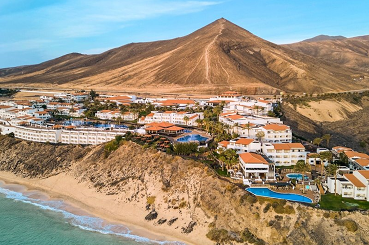 Hotelanlage mit mehreren Gebäuden und Pools auf einer Klippe am Strand vor einem großen Berg.