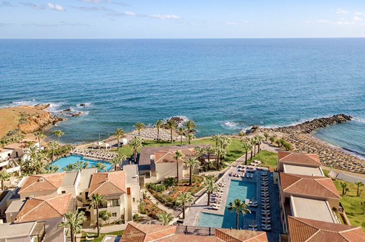 Luftaufnahme einer Hotelanlage mit mehreren Gebäuden, zwei Pools, Palmen und angrenzendem Meer.