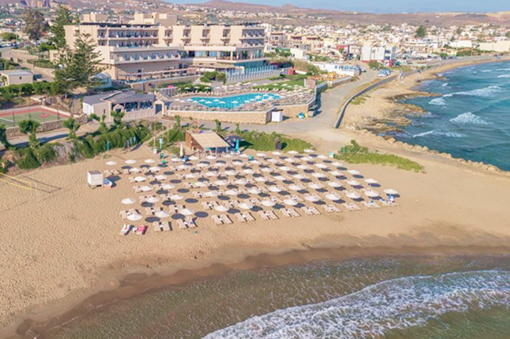 Luftaufnahme eines Strandes mit einem Hotel, umgeben von türkisblauem Wasser und sanften Wellen.