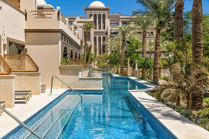 Langer Pool mit Treppen und Handläufen neben modernen Gebäuden und Palmen unter blauem Himmel Langer Pool mit Treppen und Handläufen neben modernen Gebäuden und Palmen unter blauem Himmel