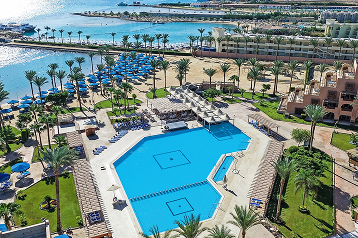 Luftaufnahme eines großen Hotelpools mit Liegen und Sonnenschirmen, umgeben von Palmen und angrenzendem Strand mit Meerblick.