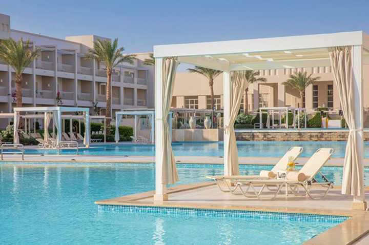 Hotelpool mit Liegestühlen und überdachten Sitzbereichen vor einem mehrstöckigen Hotelgebäude.