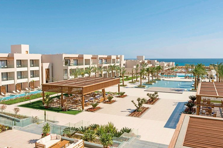 Blick auf eine moderne Hotelanlage mit mehreren zweistöckigen Gebäuden, Poolanlagen, Palmen und Sitzbereichen unter Holzpergolen am Meer.