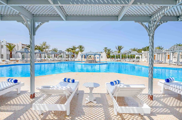 Großer Pool mit Liegen und Sonnenschirmen unter einem weißen Pavillon in einer Hotelanlage mit Palmen im Hintergrund.