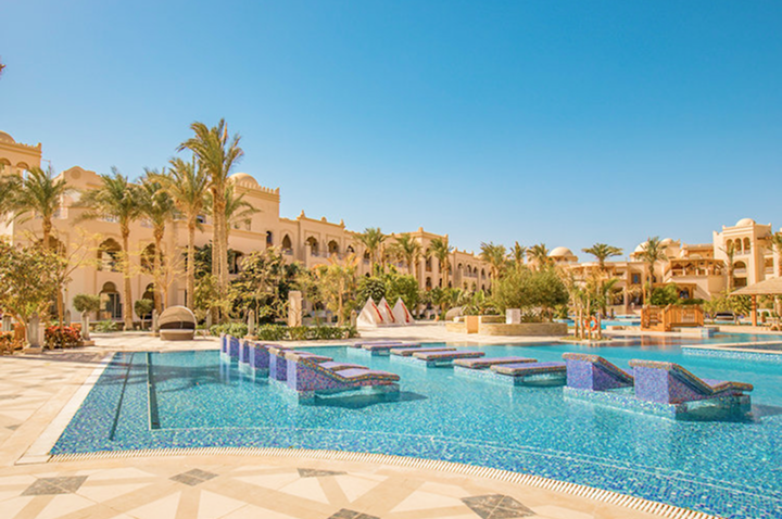 Große Hotelanlage mit mehreren Gebäuden, Palmen und Pool mit Liegeflächen im Vordergrund.