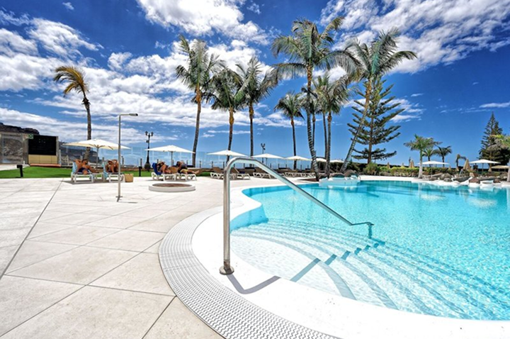 Pool mit Treppe und Edelstahlgeländer umgeben von Liegestühlen, Sonnenschirmen und Palmen unter blauem Himmel.