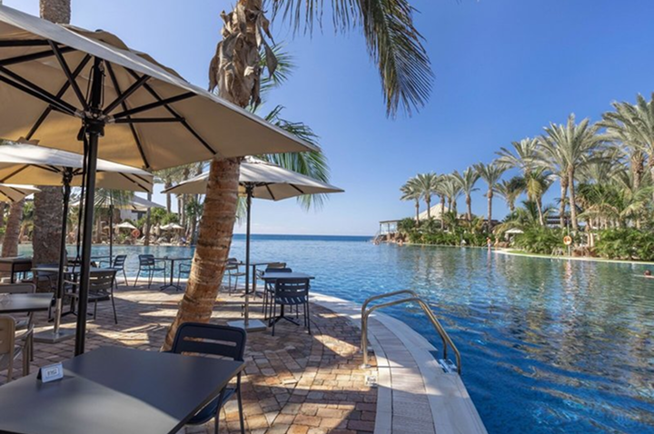 Poolbereich mit Sonnenschirmen, Tischen und Stühlen neben einem großen Schwimmbecken und Palmen im Hintergrund. Poolbereich mit Sonnenschirmen, Tischen und Stühlen neben einem großen Schwimmbecken und Palmen im Hintergrund.