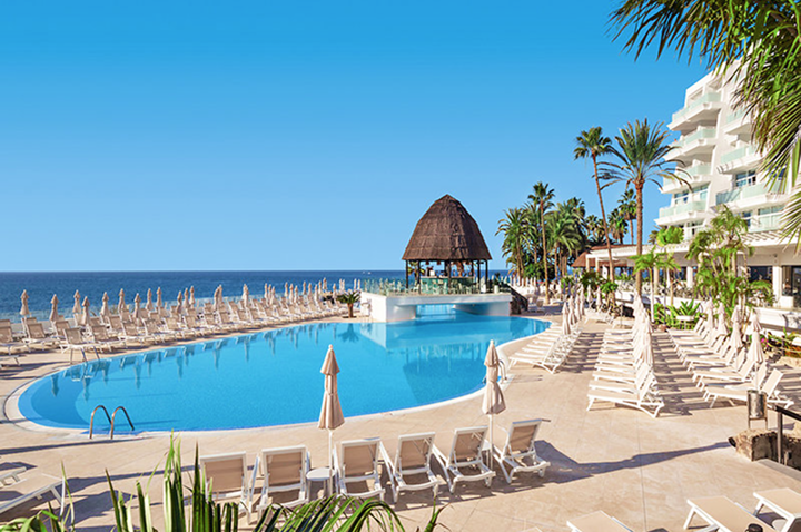 Großer runder Pool mit vielen leeren Liegestühlen und einer überdachten Poolbar vor einem Hotelgebäude am Meer.