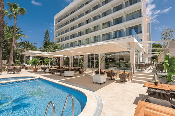 Moderner mehrstöckiger Hotelbau mit Glasfassade neben einem Pool und überdachtem Außenbereich mit Tischen und Stühlen.