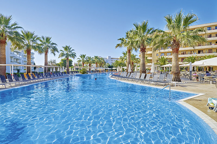 Großer Außenpool mit Palmen und Liegestühlen an beiden Seiten vor mehrstöckigen Hotelgebäuden.