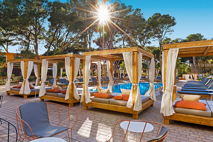 Poolbereich mit mehreren überdachten Holzliegen mit weißen Vorhängen und orangen Kissen sowie Stühlen und Tischen im Vordergrund.