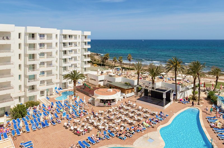 Hotelanlage mit Pool, Liegestühlen, Tischen und Stühlen auf der Terrasse, Palmen und Strand im Hintergrund.