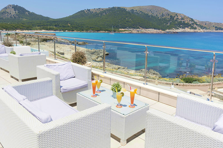 Terrasse mit weißem Rattanmöbel-Set und bunten Cocktails mit Meerblick und bewaldeten Hügeln im Hintergrund.