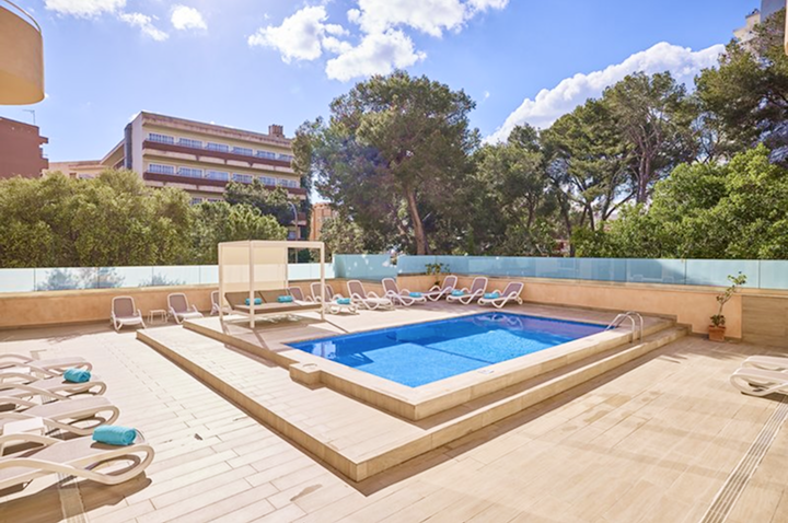 Großer Außenpool mit Liegestühlen und Sonnenschirmen vor einem mehrstöckigen Hotelgebäude und Palmen unter klarem Himmel.