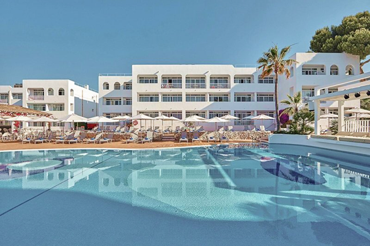 Hotelgebäude mit mehreren Stockwerken und Balkonen, davor ein großer Pool mit Liegestühlen und Sonnenschirmen, Palmen am Rand.