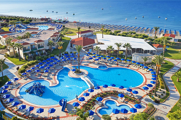 Luftaufnahme eines Resorts mit mehreren Pools, Sonnenschirmen, Gebäuden mit einer Bucht mit Sandstrand und Felsen.