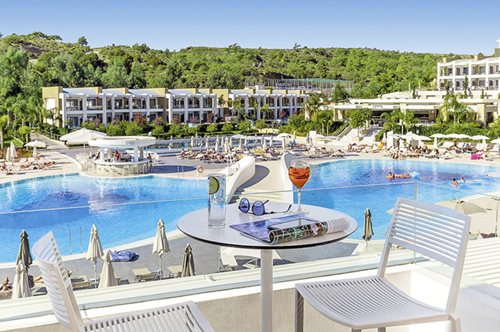 Blick von einer Terrasse mit Tisch und zwei Stühlen auf eine große Poollandschaft vor einem modernen Hotelkomplex mit mehreren Gebäuden und viel Grün im Hintergrund.