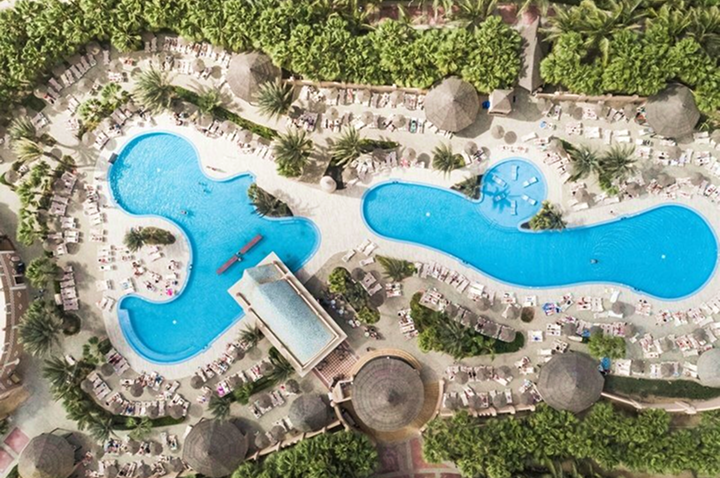 Luftaufnahme eines großen, organisch geformten Schwimmbeckens mit Liegestühlen und Sonnenschirmen umgeben von dichtem Grün.