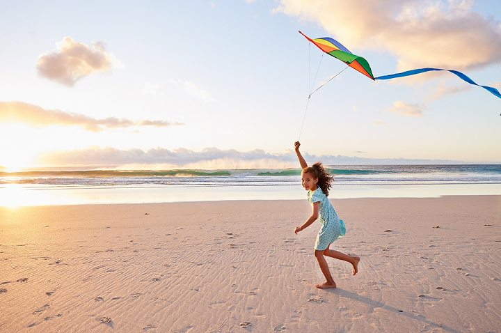 Kind in blau-weiß gepunktetem Kleid läuft barfuß am Strand und lässt einen bunten Drachen steigen.