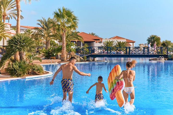 Drei Personen in Badesachen laufen am Rand eines großen Pools, eine Frau trägt eine bunte Schwimmliege in Form eines Eis am Stiel.